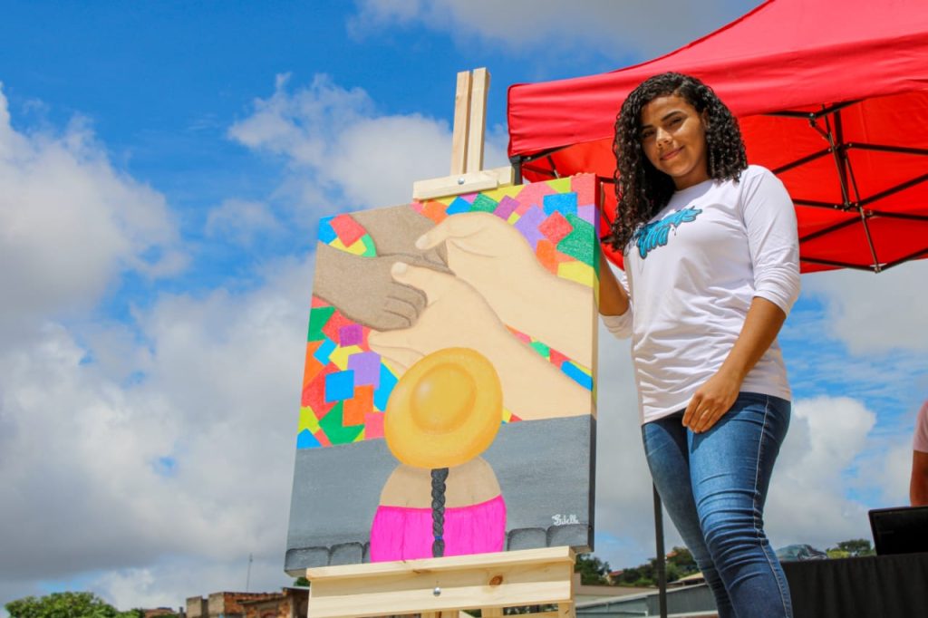 A jovem Sibelle está ao lado de um quadro pintado por ela, em evento da organização parceira.